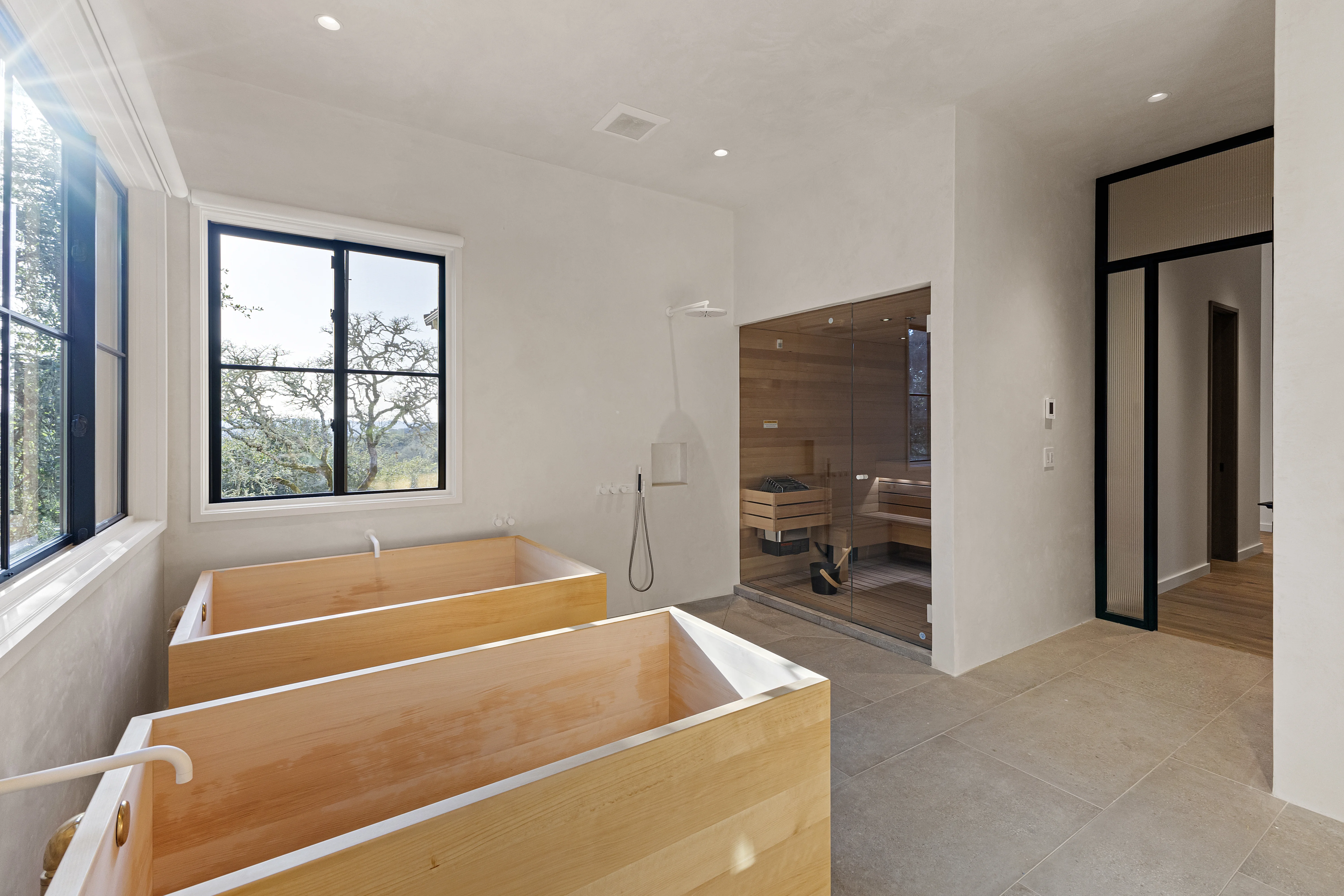 Japanese-style soaking tub and glass-enclosed cedar sauna in a minimalist white bathroom with garden views