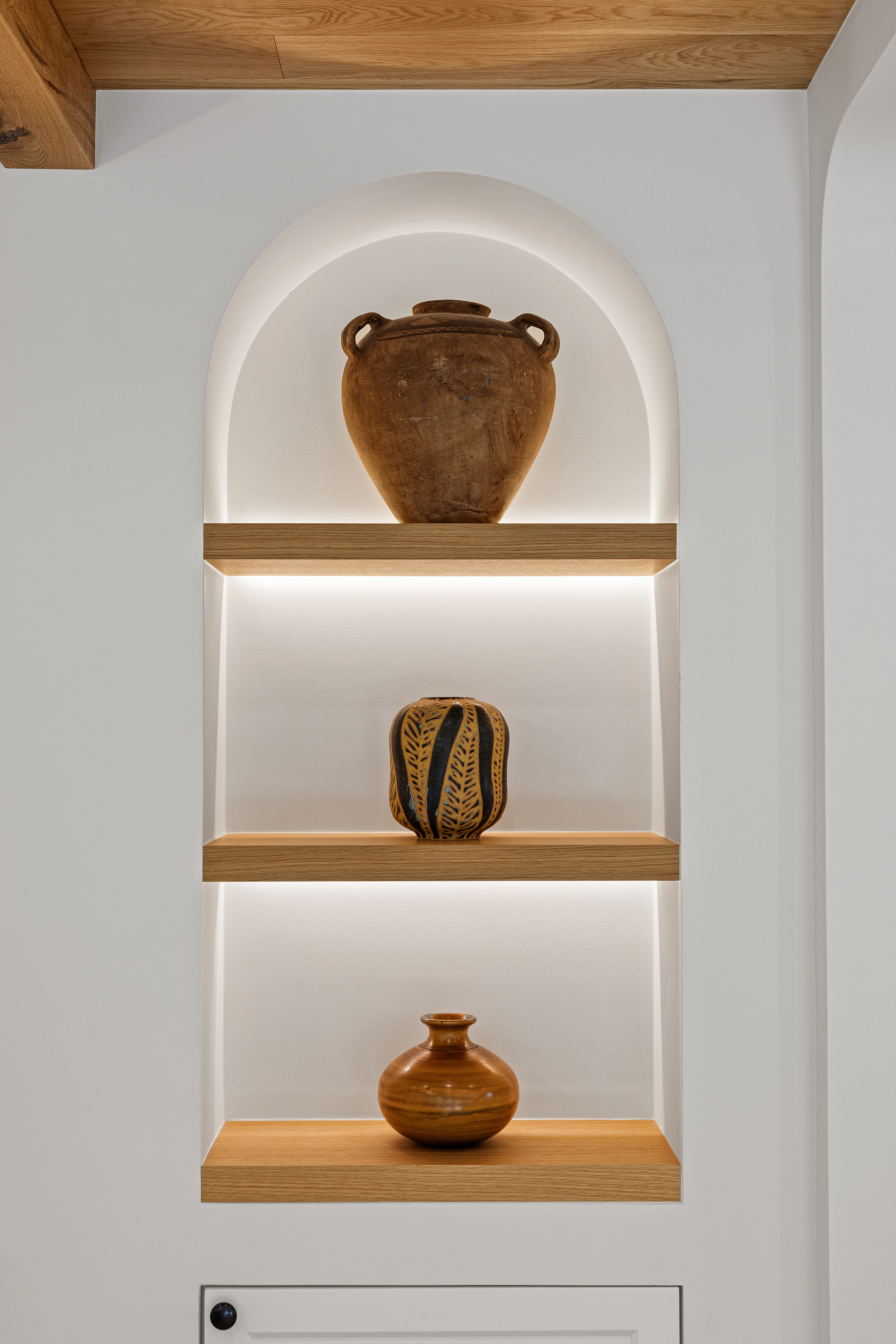 Arched alcove niche with backlit oak shelves displaying handcrafted ceramic vessels